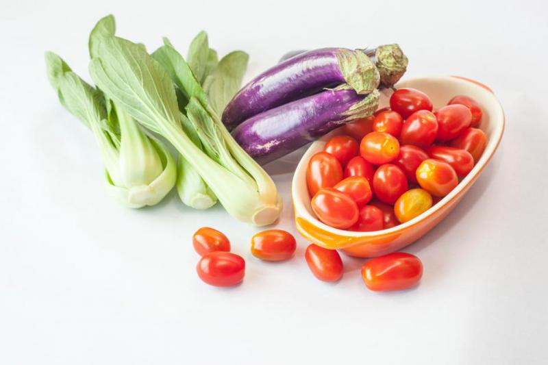 There is a spread of three types of fruits and vegetables. There is a small, long bowl of grape tomatoes with some of them having spilled out in front of  the bowl. There are three grapefruits leaning on the left side of the bowl. To the left of the grapefruits are three stalks of bok choy.