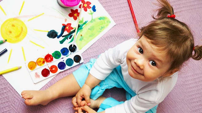 A young girl  is sitting on the ground with a water color paint set and a partially completed painting of flowers growing under a yellow sun. 