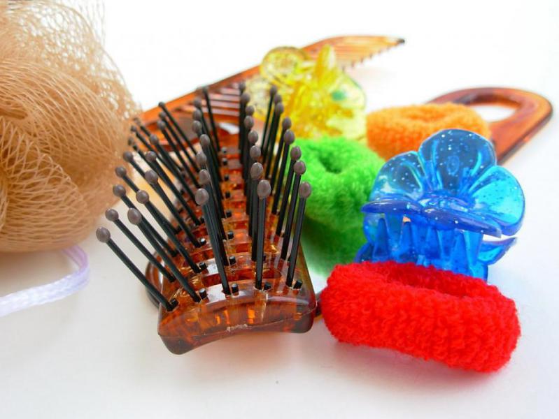 A brush is sitting next to a pile of hair accessories. To the right of the brush there is a red hair tie, a blue hair clip, a green hair tie, an orange hair tie, two yellow hair clips, and a brown comb. To the left of the brush is a light brown loofa.