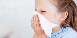 A child wearing a blue shirt is holding a tissue to cover her nose and mouth while sneezing.
