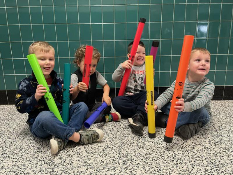 Four young children are seen holding two Boomwhackers each. The children are sitting on the floor of a school hallway. The Boomwhackers are different shades of red, green, and yellow.