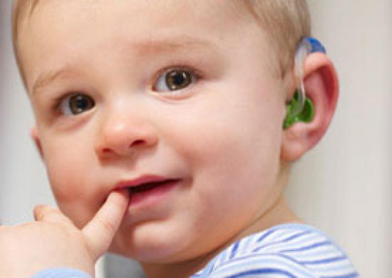 A young child with a hearing aid. Their finger is in their mouth and they are looking almost directly at the viewer of the picture.