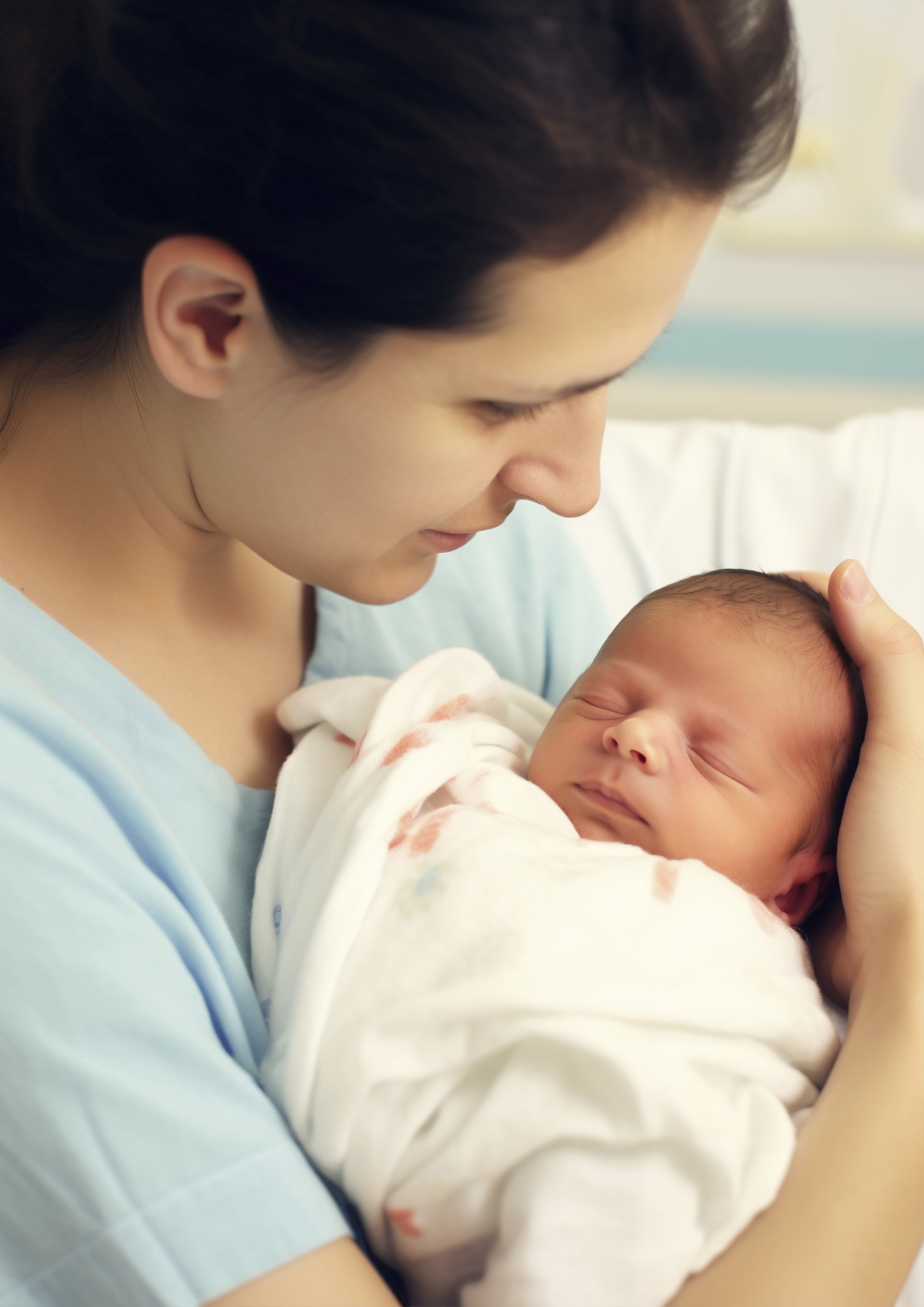 Two parents looking at their newborn baby together in front of a window. The two parents are embracing one another as the mother holds their baby. The baby is swaddled in a white polka dot blanket.