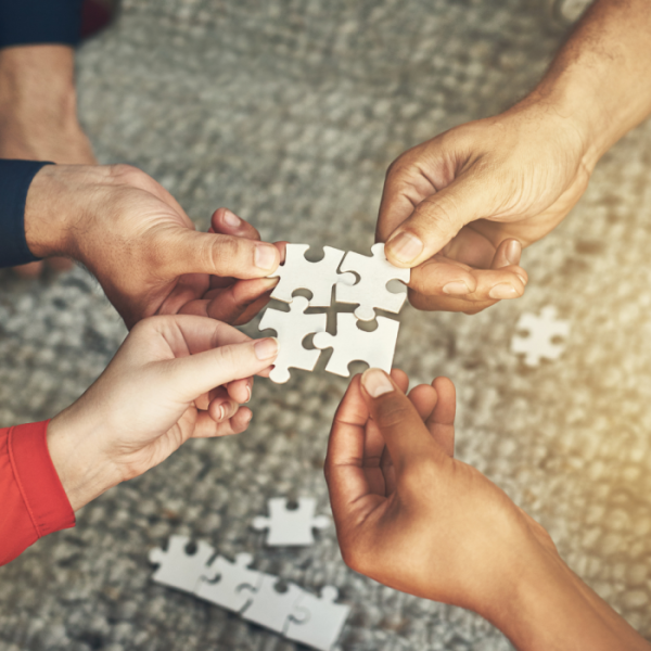 Four people are holding out a puzzle piece. Each piece is being put together to form a square in the middle of everyone's hands.
