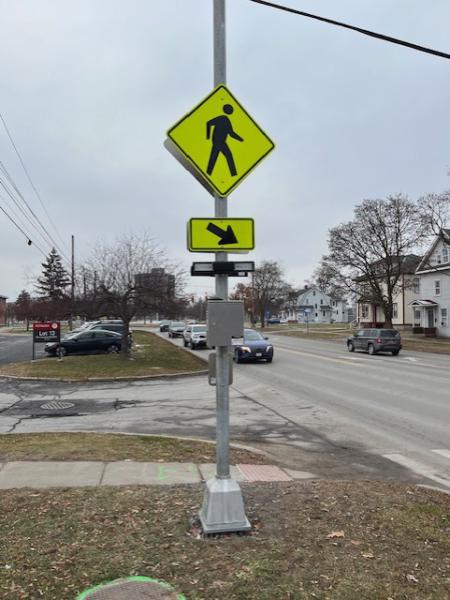 A yellow crosswalk sign depicting a person walking with an arrow pointing towards the road. The sign is equipped with a button that turns on flashing yellow lights to safely alert drivers that a person is using the crosswalk.